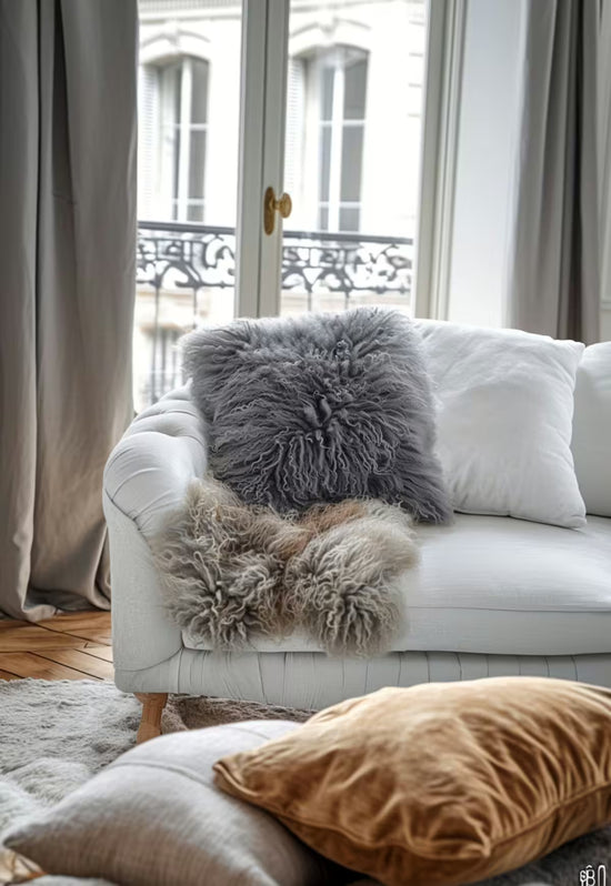 Lammfellkissen in Grau als Detail auf dem Sofa, neben Kissen und vor dem Fenster.