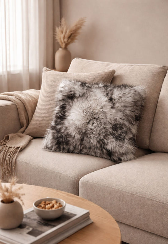 Graues Lammfellkissen auf hellem Sofa mit weiteren Kissen und Decke im Wohnraum.