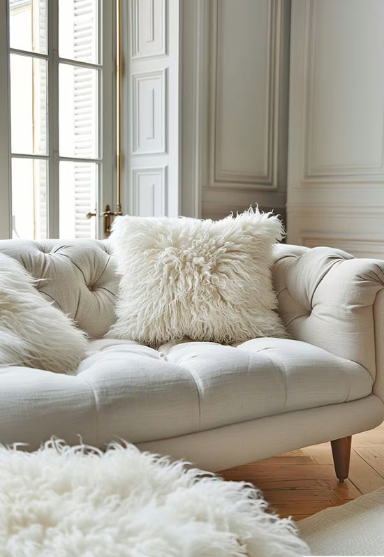 Weißes Lammfellkissen als Detail auf einem gepolsterten Sofa vor hohen Fenstern und hellem Parkett.