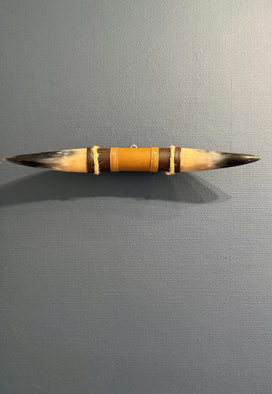 Wanddekoration aus Naturhorn mit Leder-Holzhalterung an grauer Wand, frontal ausgerichtet.